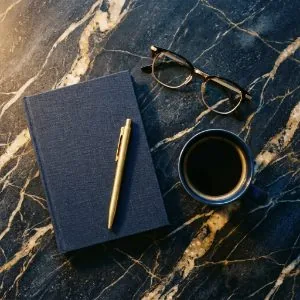 A navy linen notebook, brass pen, reading glasses and black coffee on dark marble